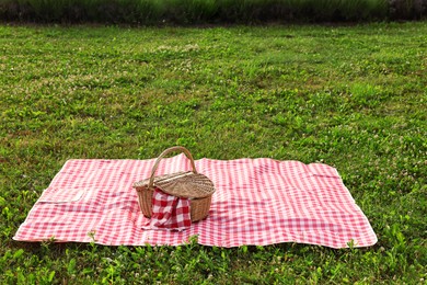 Photo of Picnic wicker basket with napkin and red checkered blanket on green grass outdoors