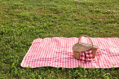 Photo of Picnic wicker basket with napkin and red checkered blanket on green grass outdoors