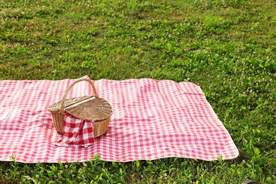 Photo of Picnic wicker basket with napkin and red checkered blanket on green grass outdoors