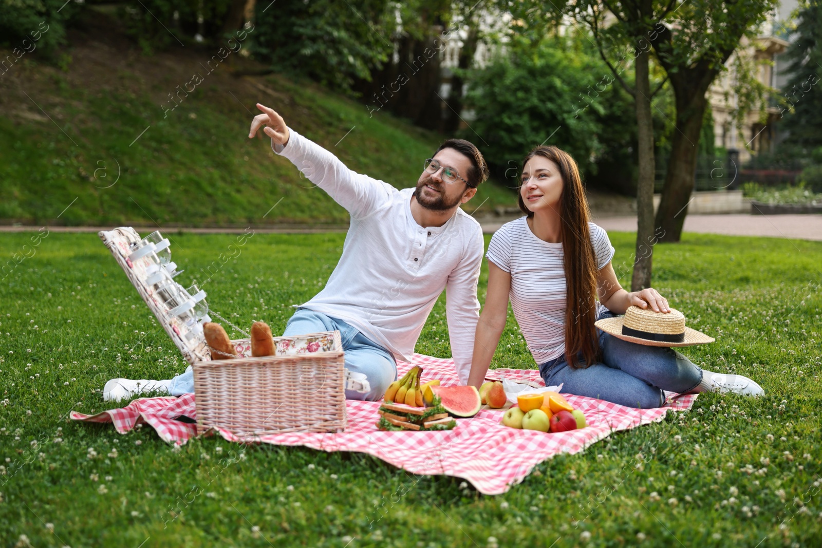 Photo of Romantic picnic. Smiling man pointing at something to his girlfriend on green grass outdoors