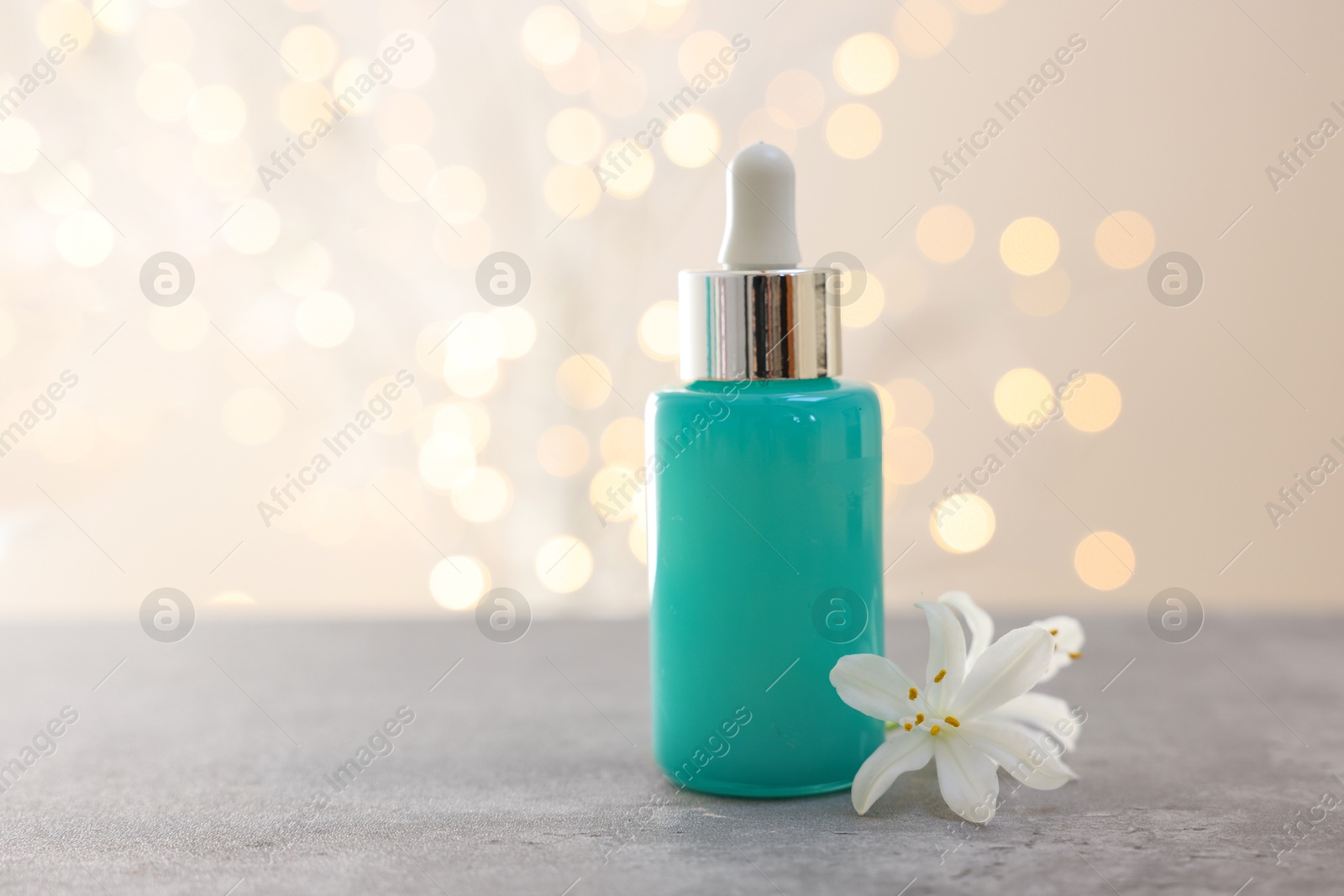 Photo of Cosmetic product in bottle and beautiful jasmine flower on grey table against beige background with blurred lights, closeup