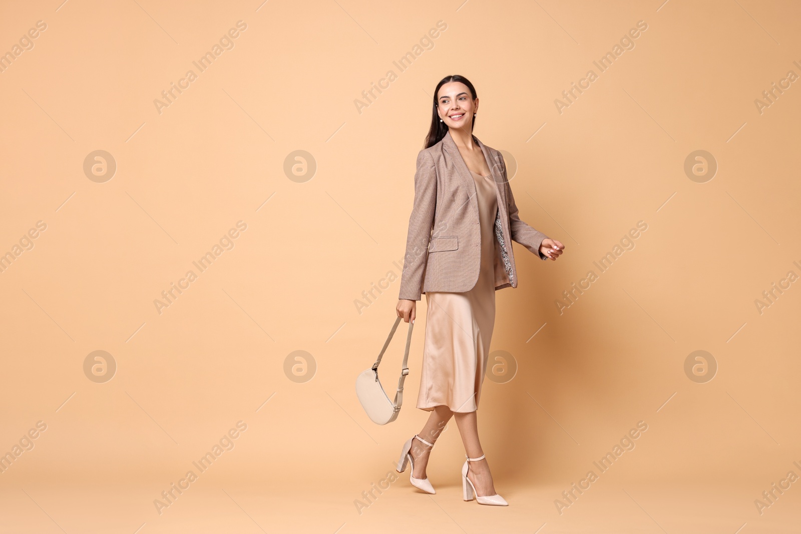 Photo of Beautiful woman in stylish jacket with bag walking on pale orange background