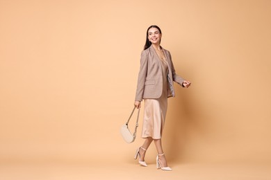 Beautiful woman in stylish jacket with bag walking on pale orange background