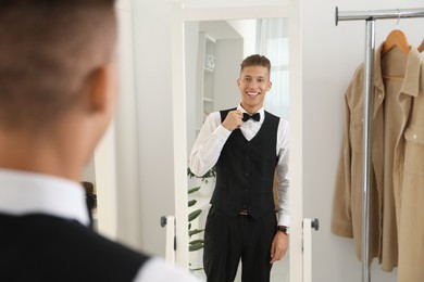 Handsome man adjusting bow tie near mirror indoors