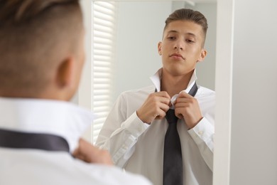 Photo of Handsome man adjusting necktie near mirror indoors