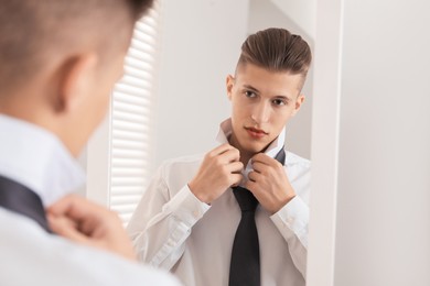 Handsome man adjusting necktie near mirror indoors