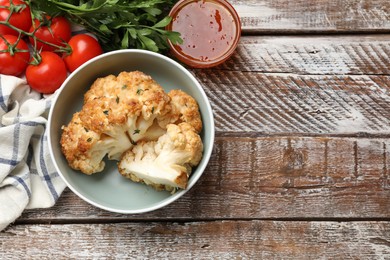 Tasty baked cauliflower in bowl, sauce, tomato and parsley on wooden table, flat lay. Space for text