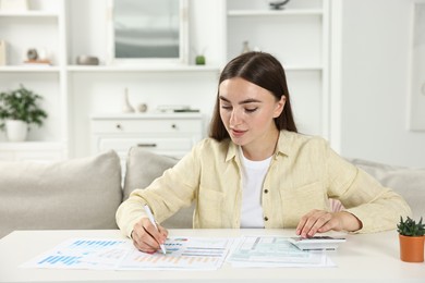 Photo of Budget planning. Beautiful young woman with papers using calculator at white table indoors