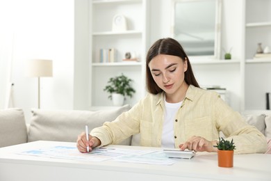 Photo of Budget planning. Beautiful young woman with papers using calculator at white table indoors