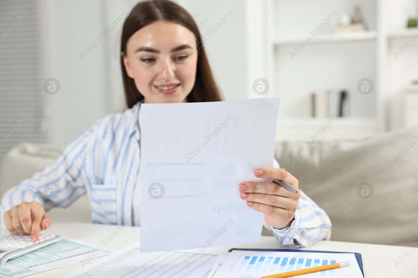 Photo of Budget planning. Beautiful young woman with papers using calculator at white table indoors, selective focus