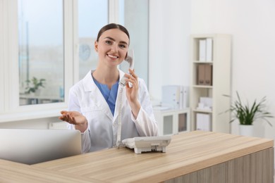 Professional receptionist talking on phone at wooden desk in hospital