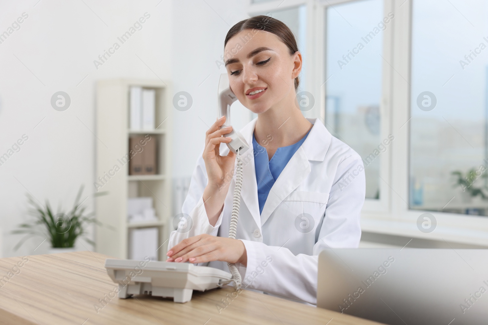 Photo of Professional receptionist talking on phone at wooden desk in hospital