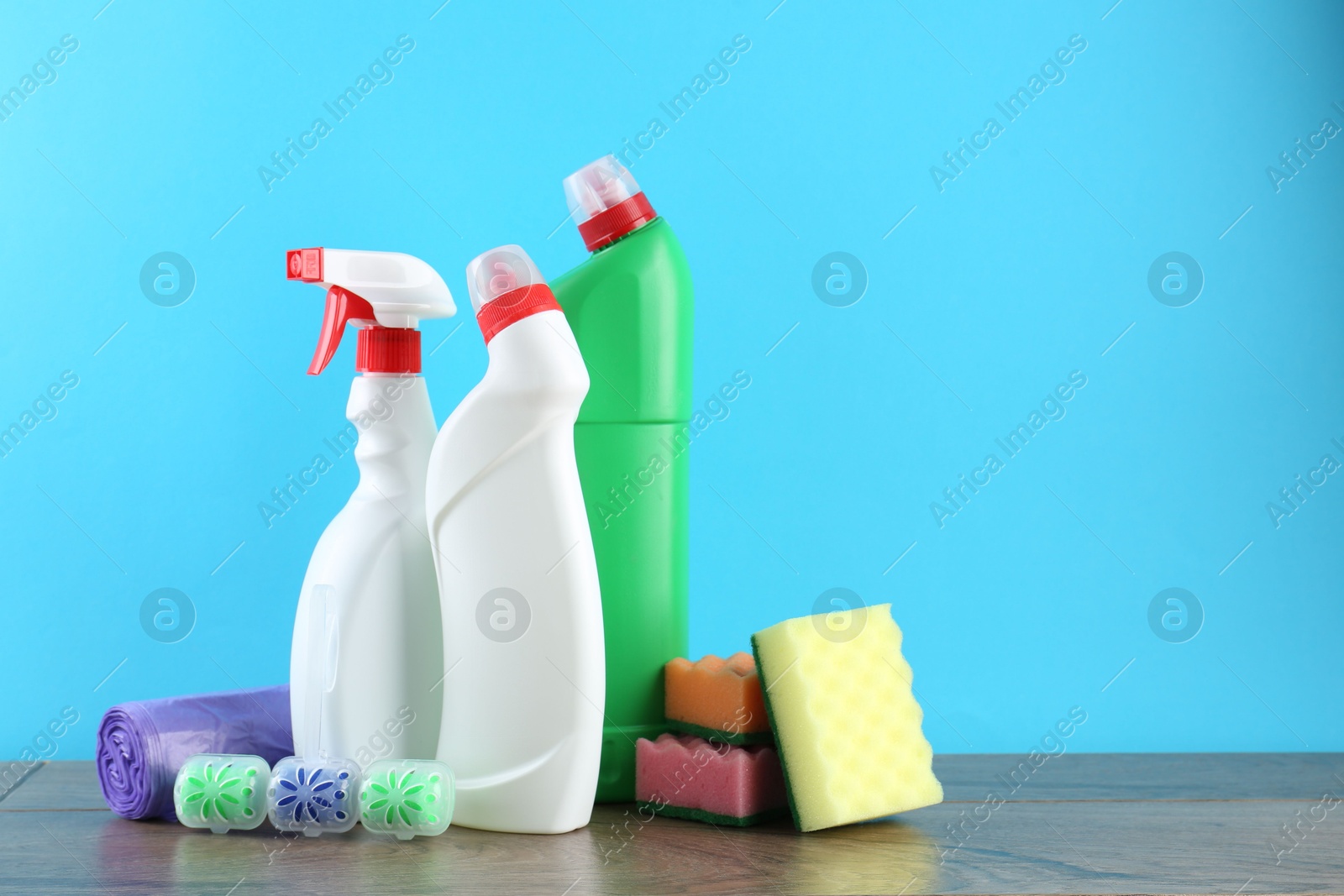 Photo of Different toilet cleaners, sponges and trash bags on wooden table against light blue background