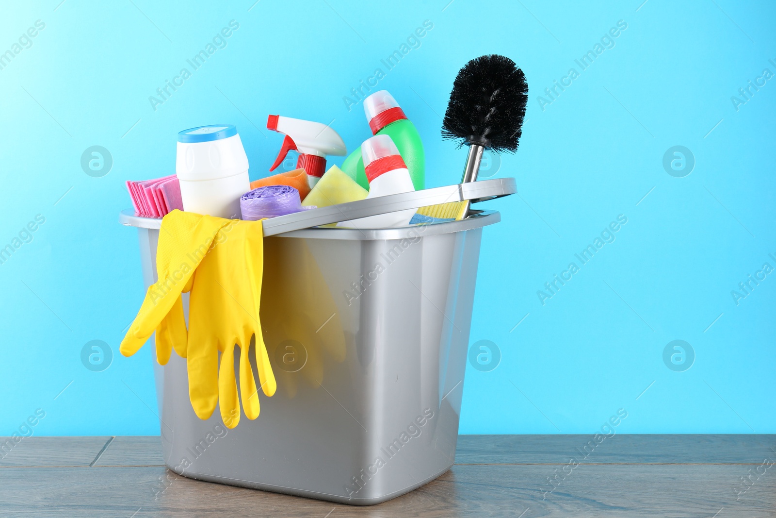 Photo of Bucket with different toilet cleaners, rags, rubber gloves and trash bags on wooden table against light blue background