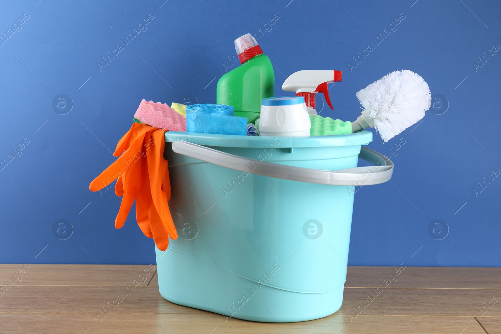 Photo of Bucket with different toilet cleaners, sponges, rubber gloves and trash bags on wooden table against blue background