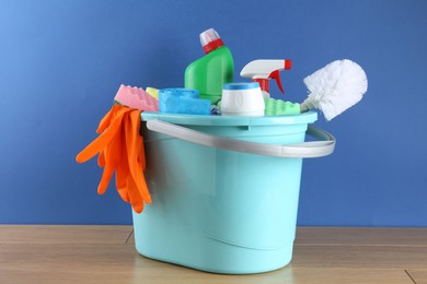 Photo of Bucket with different toilet cleaners, sponges, rubber gloves and trash bags on wooden table against blue background