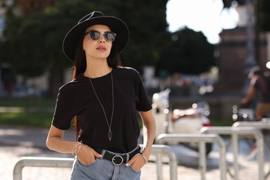 Photo of Young woman in stylish black hat and sunglasses on city street, space for text