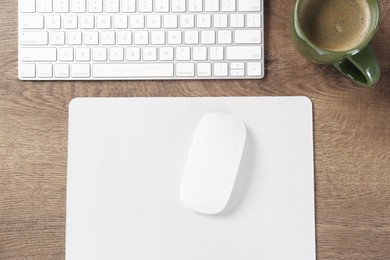 Photo of Computer mouse with mousepad, keyboard and cup of coffee on wooden table, flat lay