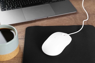 Photo of Computer mouse with mousepad, cup of coffee and laptop on wooden table