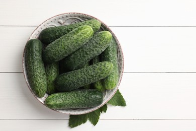 Photo of Fresh cucumbers in bowl and green leaves on white wooden table, top view. Space for text