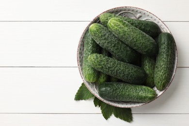 Photo of Fresh cucumbers in bowl and green leaves on white wooden table, top view. Space for text