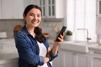 Smiling woman with smartphone in kitchen. Space for text