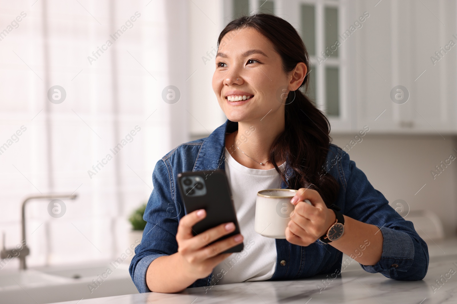 Photo of Smiling woman with smartphone drinking coffee at table in kitchen