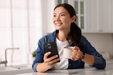 Smiling woman with smartphone drinking coffee at table in kitchen