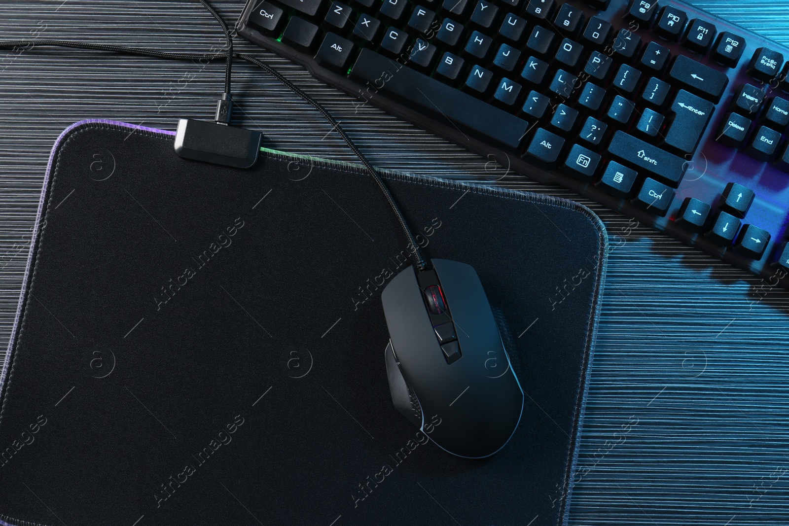 Photo of Computer mouse, mousepad and keyboard on dark wooden table in neon lights, flat lay