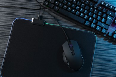 Photo of Computer mouse, mousepad and keyboard on dark wooden table in neon lights, flat lay