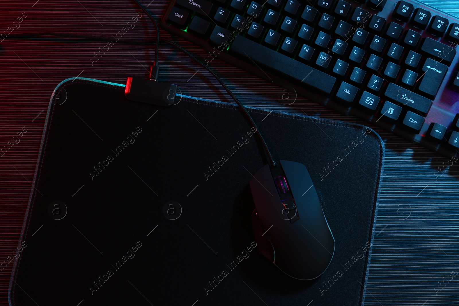 Photo of Computer mouse, mousepad and keyboard on dark wooden table in neon lights, flat lay