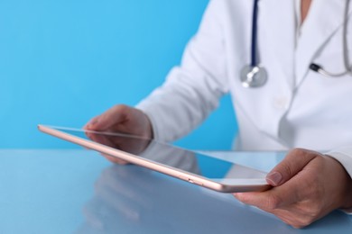 Doctor with tablet at table against light blue background, closeup view