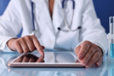 Doctor with tablet at table against blue background, closeup view