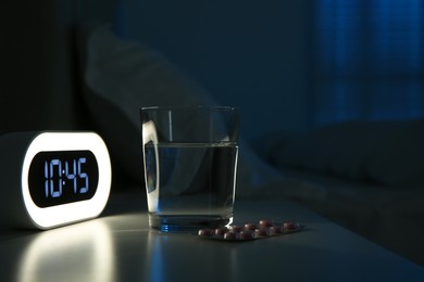 Photo of Insomnia treatment. Glass of water, pills and alarm clock on bedside table in bedroom at night