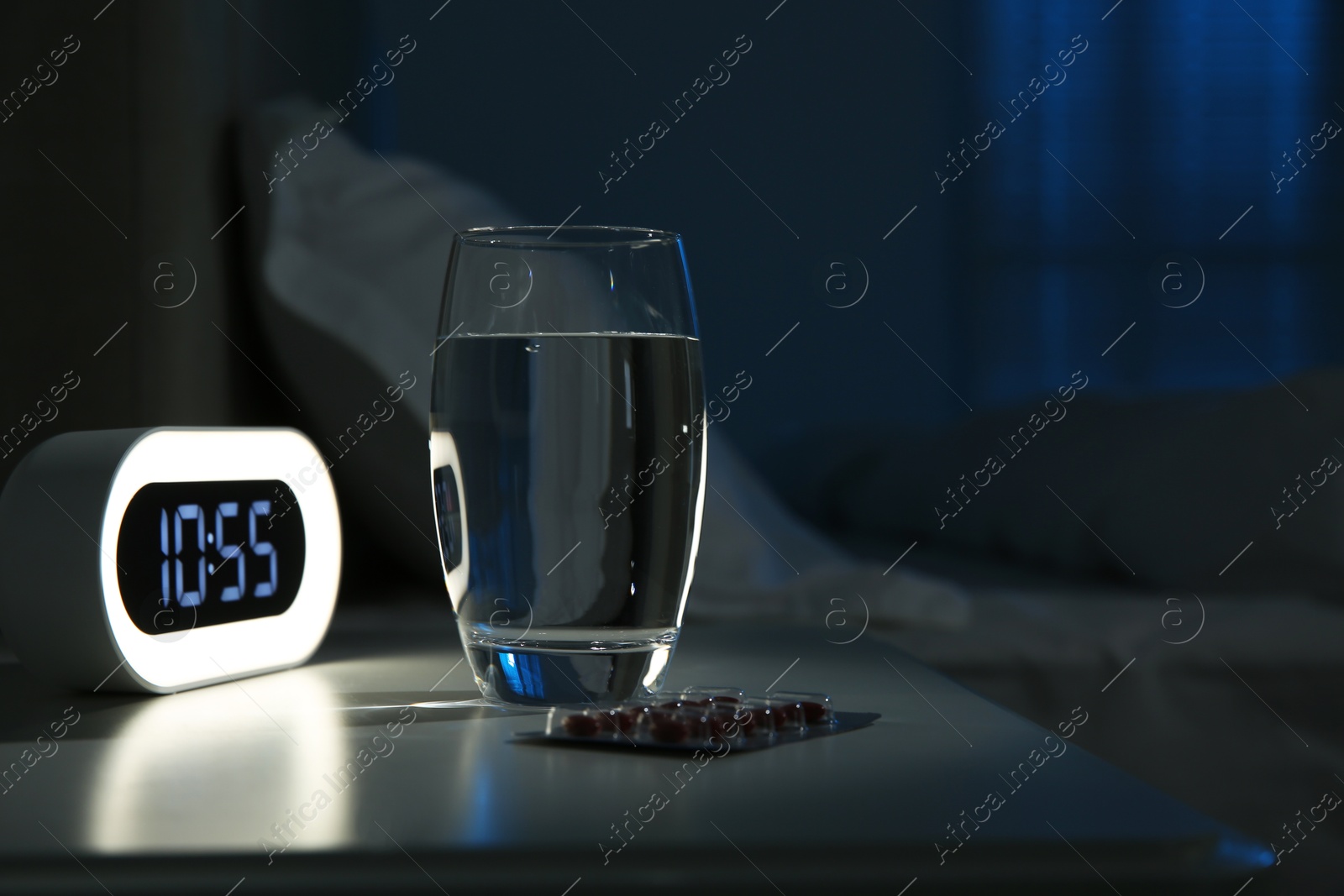 Photo of Insomnia treatment. Alarm clock, glass of water and pills on bedside table in bedroom at night