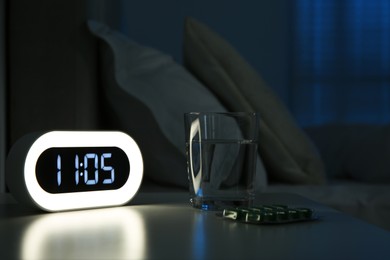 Insomnia treatment. Glass of water, pills and alarm clock on bedside table in bedroom at night