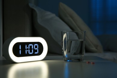 Insomnia treatment. Glass of water, pills and alarm clock on bedside table in bedroom at night