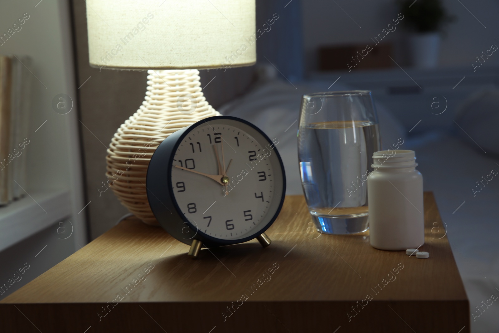 Photo of Insomnia treatment. Glass of water, pills and alarm clock on bedside table in bedroom at night