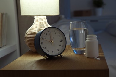 Insomnia treatment. Glass of water, pills and alarm clock on bedside table in bedroom at night