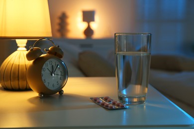 Insomnia treatment. Glass of water, pills and alarm clock on bedside table in bedroom at night