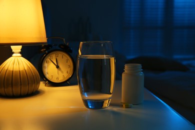 Photo of Insomnia treatment. Glass of water, pills and alarm clock on bedside table in bedroom at night