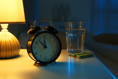 Photo of Insomnia treatment. Alarm clock, glass of water and pills on bedside table in bedroom at night