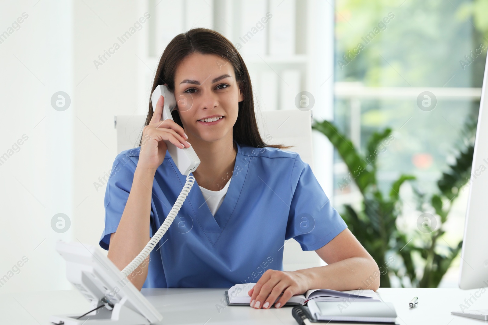 Photo of Smiling nurse working with clients by telephone at table in hospital office