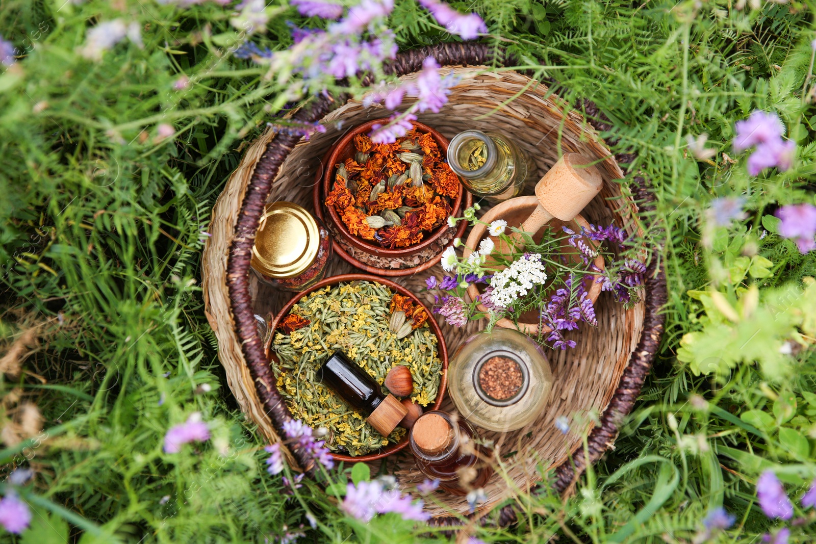 Photo of Different ingredients for tincture, mortar and pestle in wicker basket on green grass outdoors, top view