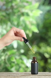 Photo of Woman dripping tincture from pipette into bottle at wooden table against blurred green background, closeup