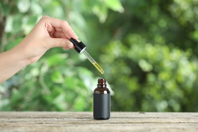 Photo of Woman dripping tincture from pipette into bottle at wooden table against blurred green background, closeup