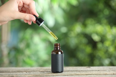 Photo of Woman dripping tincture from pipette into bottle at wooden table against blurred green background, closeup