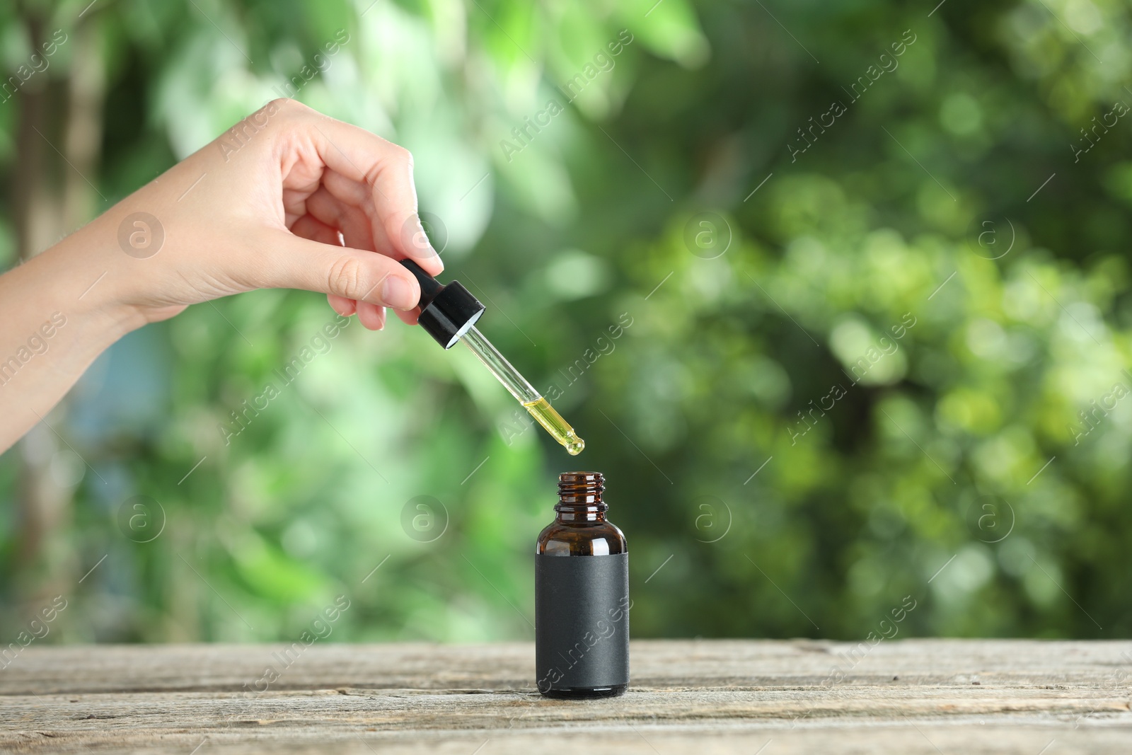 Photo of Woman dripping tincture from pipette into bottle at wooden table against blurred green background, closeup