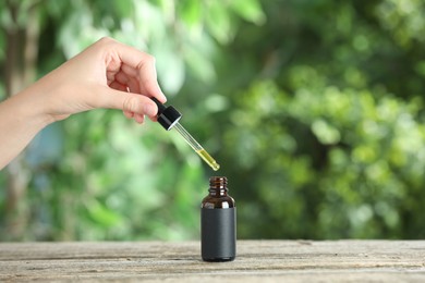 Photo of Woman dripping tincture from pipette into bottle at wooden table against blurred green background, closeup