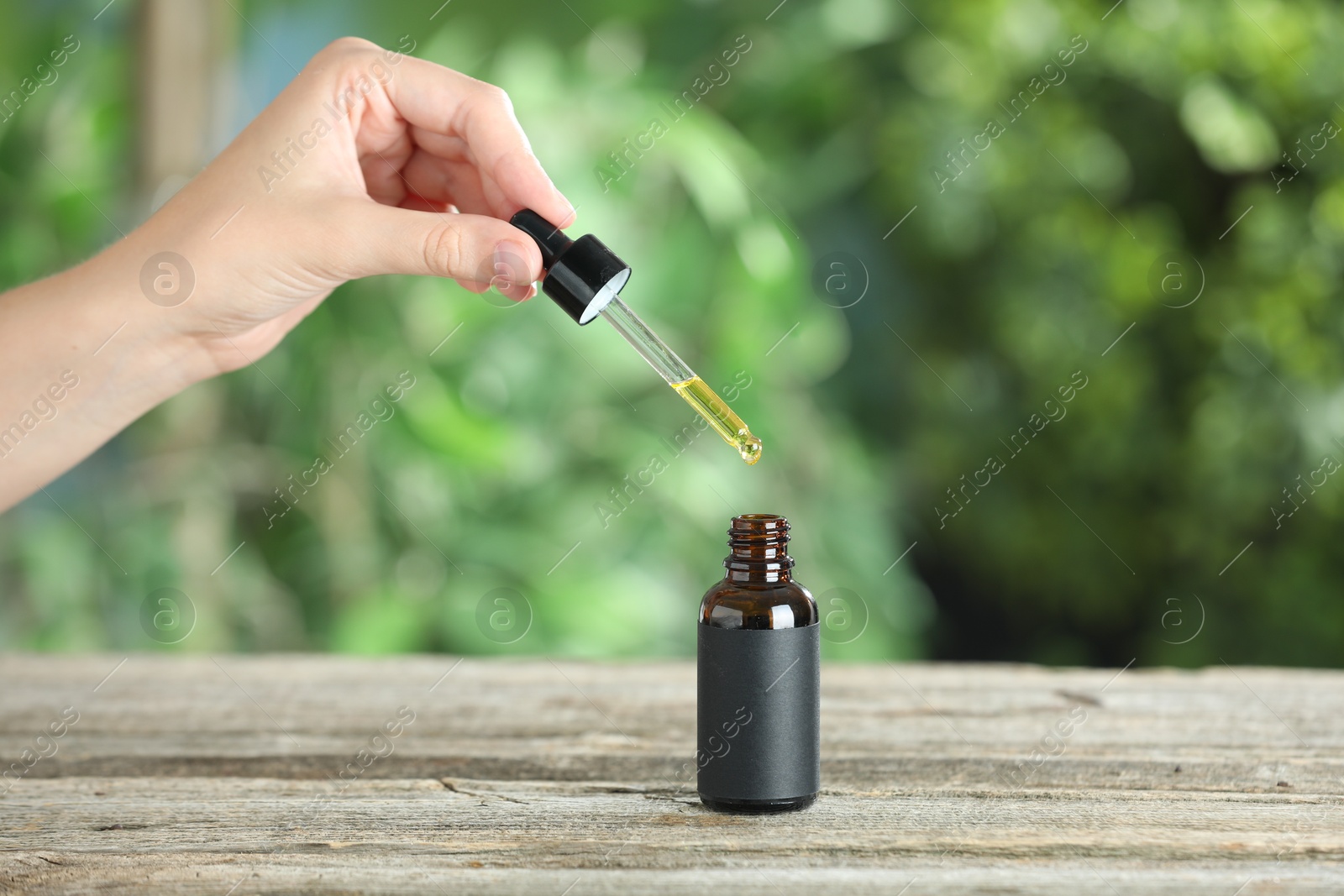 Photo of Woman dripping tincture from pipette into bottle at wooden table against blurred green background, closeup